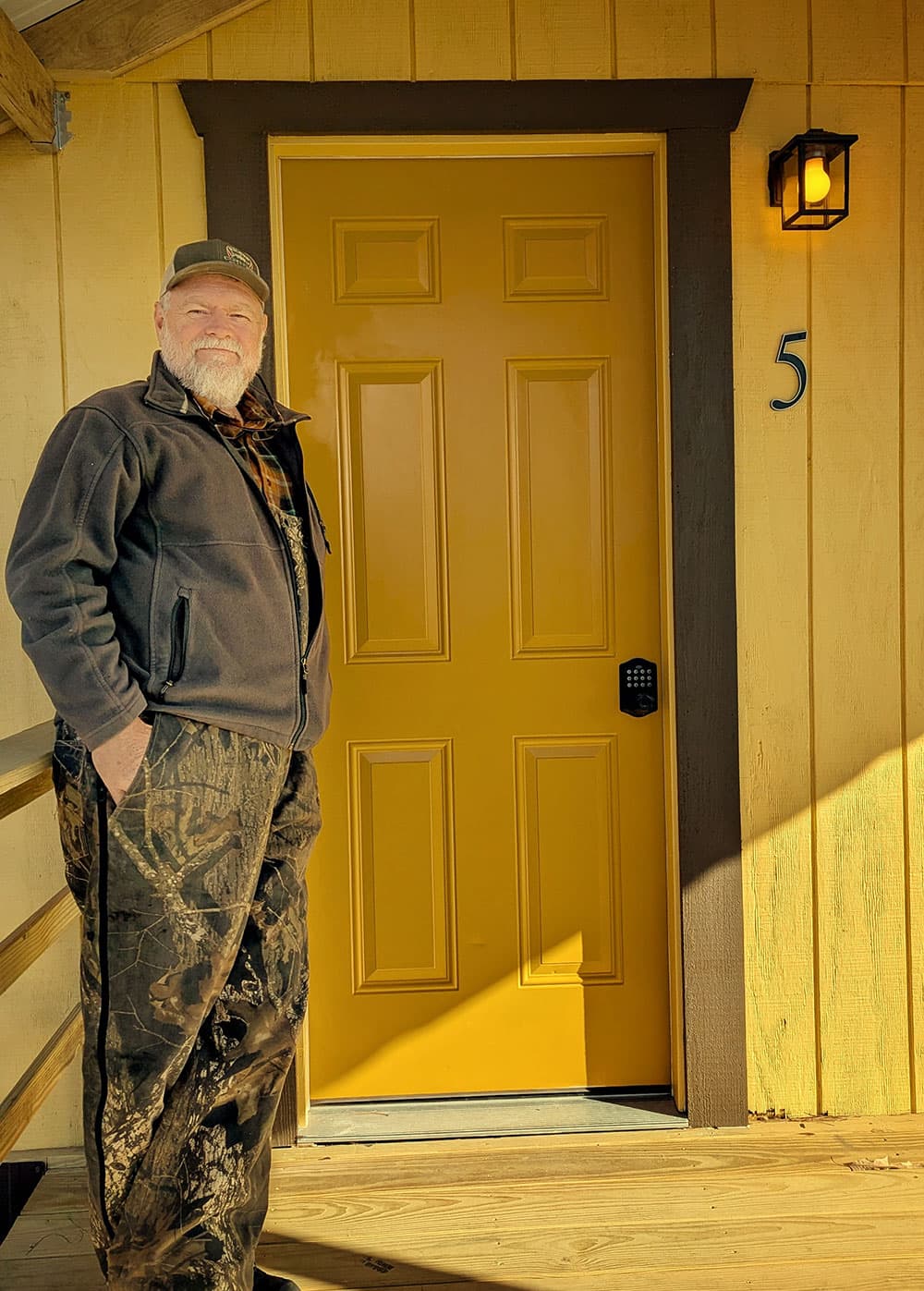 A man stands beside a bright yellow door on a wooden porch.