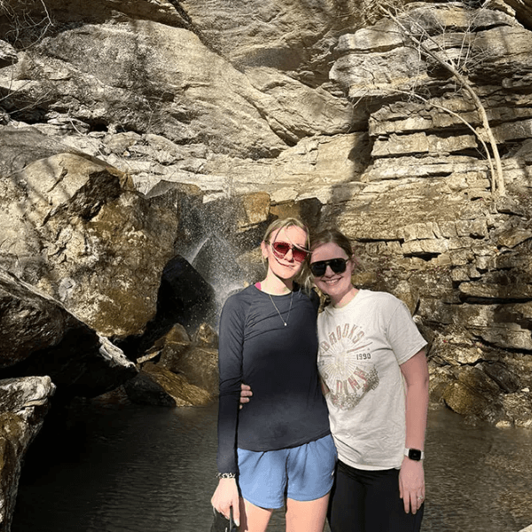 Two friends pose together in front of a waterfall and rocky backdrop.