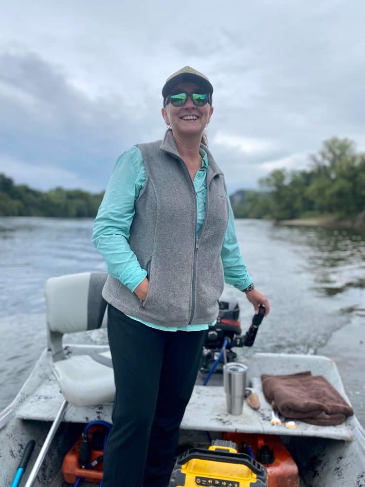 A woman smiles while standing in a small boat on a river, wearing sunglasses and a vest.