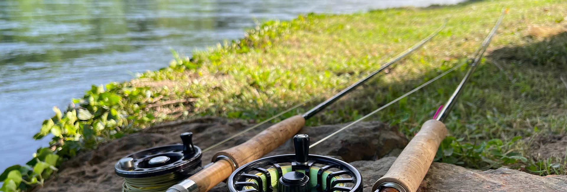 Two fishing rods and reels resting on the grass by a riverbank.