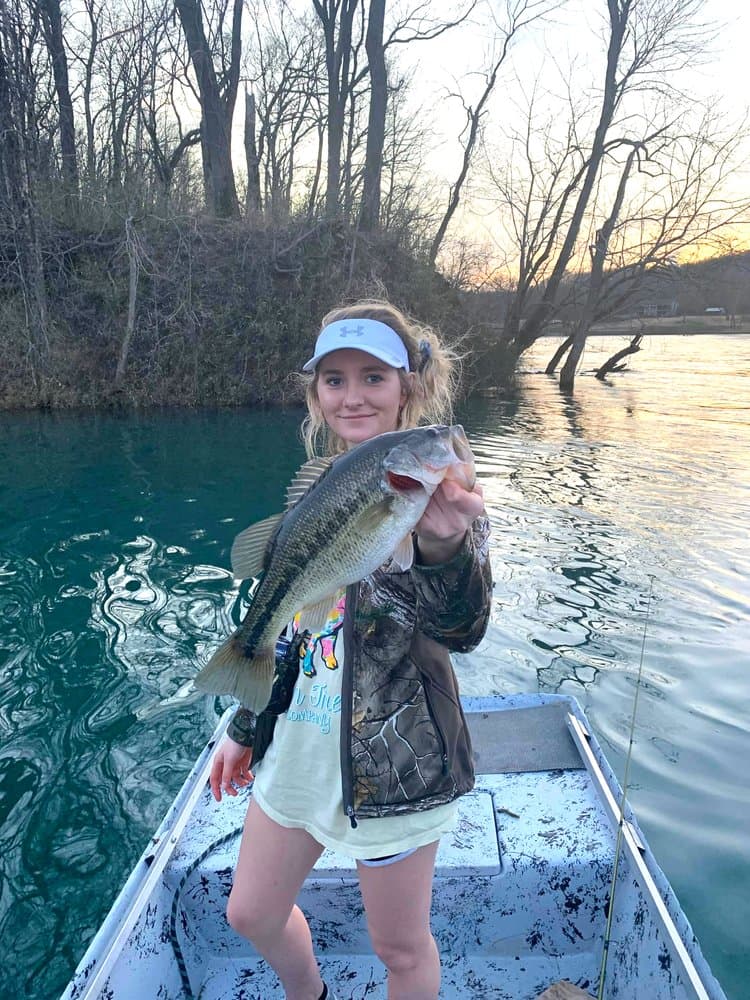 A girl in a boat holds up a large bass fish with a smile, surrounded by trees and water at sunset.