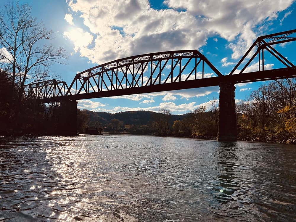 A silhouette of a bridge spans a river under a bright sky with scattered clouds.