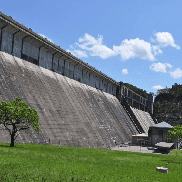A large concrete dam stands against a blue sky with scattered clouds, surrounded by green grass and trees.