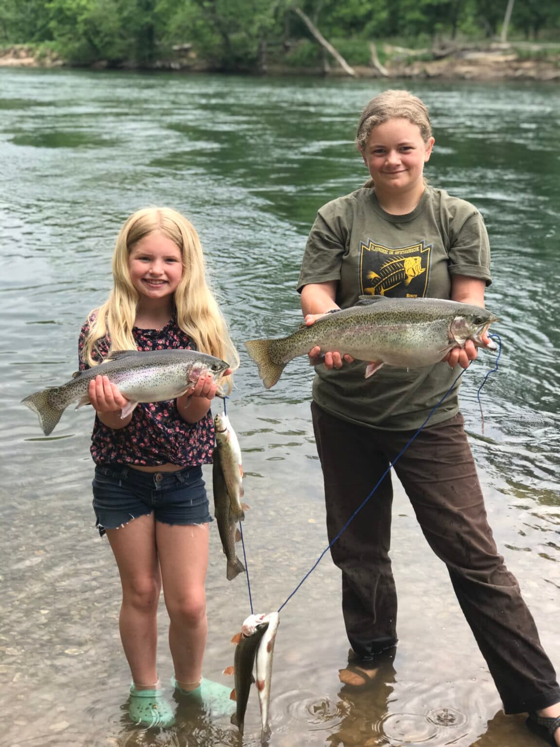 Two girls stand in shallow water, smiling and holding large fish.
