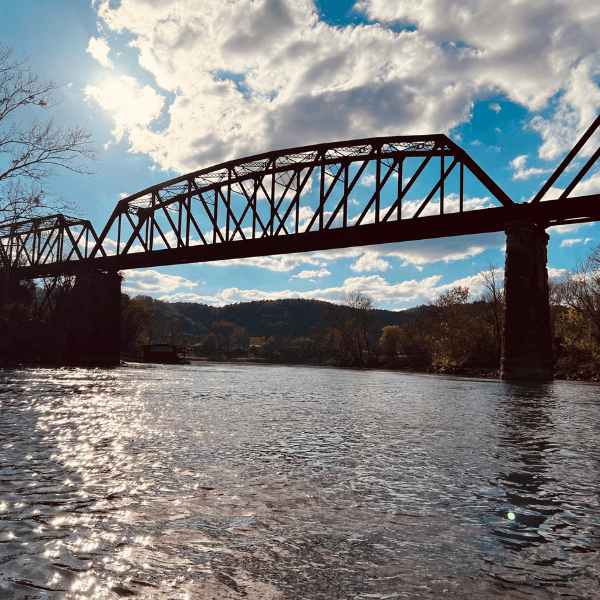 A silhouetted railroad bridge spans a reflective river under a blue sky filled with clouds.