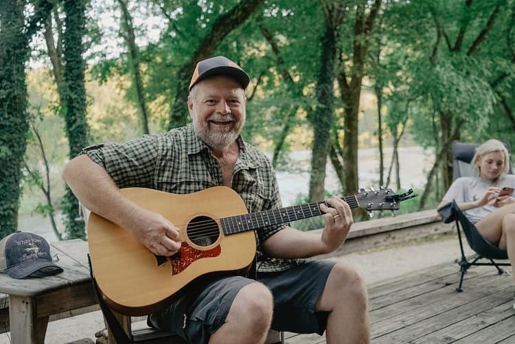 A smiling man plays guitar outdoors while another person relaxes behind him.
